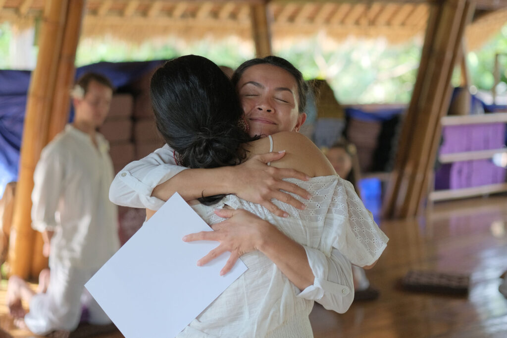 Two women in white clothes hugging during the yoga teacher training closing ceremony
