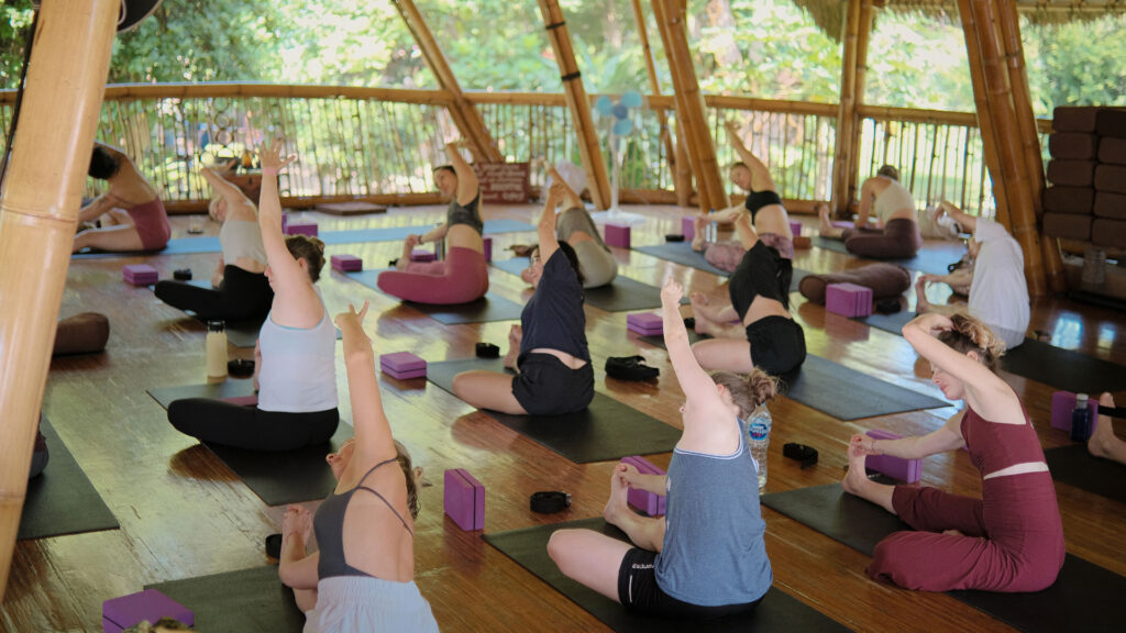 Group of yoga students in a seated side stretch during class at Power of Now Oasis’ bamboo studio in Bali