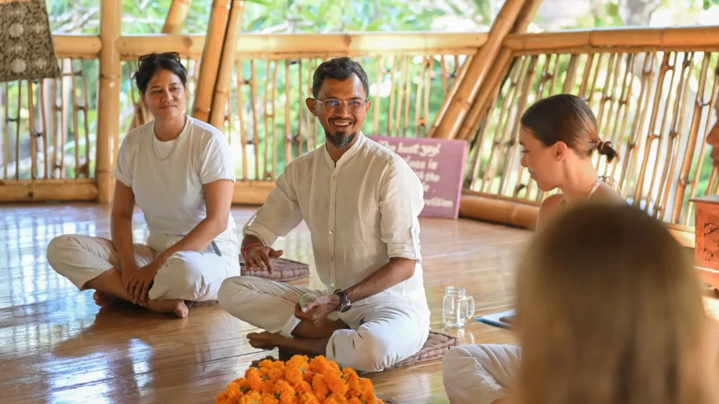 Krishna, head teacher at Power of Now Oasis, sitting cross‑legged and smiling during the 200‑Hour Yoga Teacher Training closing ceremony in the bamboo shala in Sanur, Bali