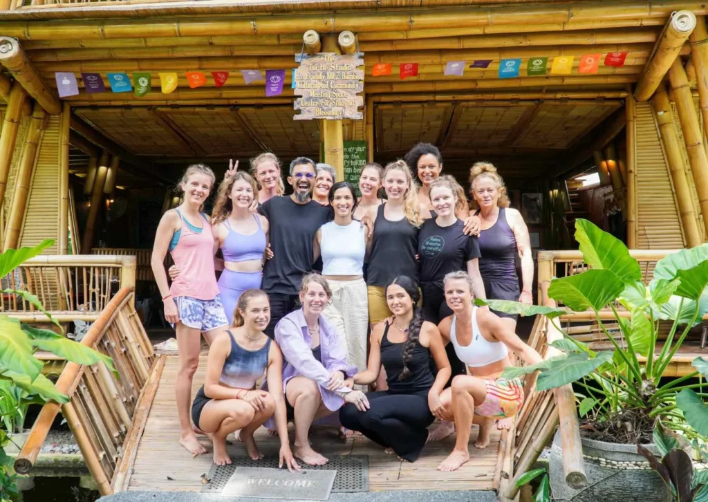 Group of Yoga Teacher Training students and teachers smiling together outside the bamboo shala at Power of Now Oasis in Sanur, Bali