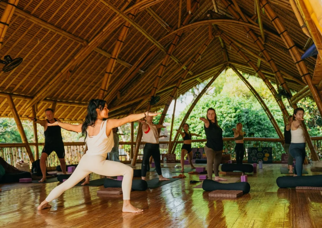 Yoga class in Bali with students practicing warrior pose in a bamboo shala surrounded by greenery