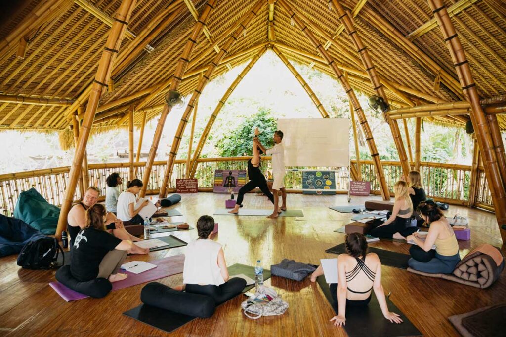 Yoga teacher adjusts a student in a warrior pose during a theory class at Power of Now Oasis in Bali